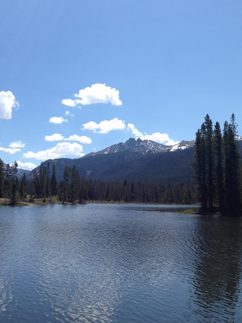 A lake, surrounded by pine trees with a rocky moutain with spots of snow in the background
