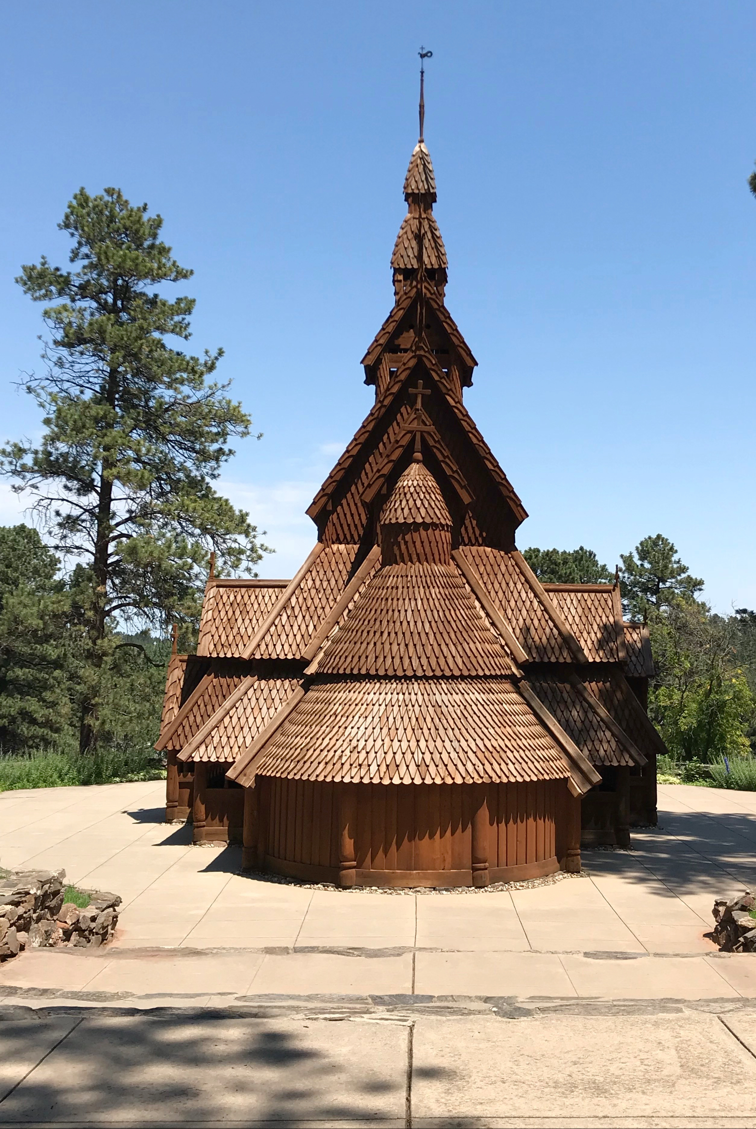 A wooden church sits on a concrete patio surrounded by pine trees, all under a clear, blue sky