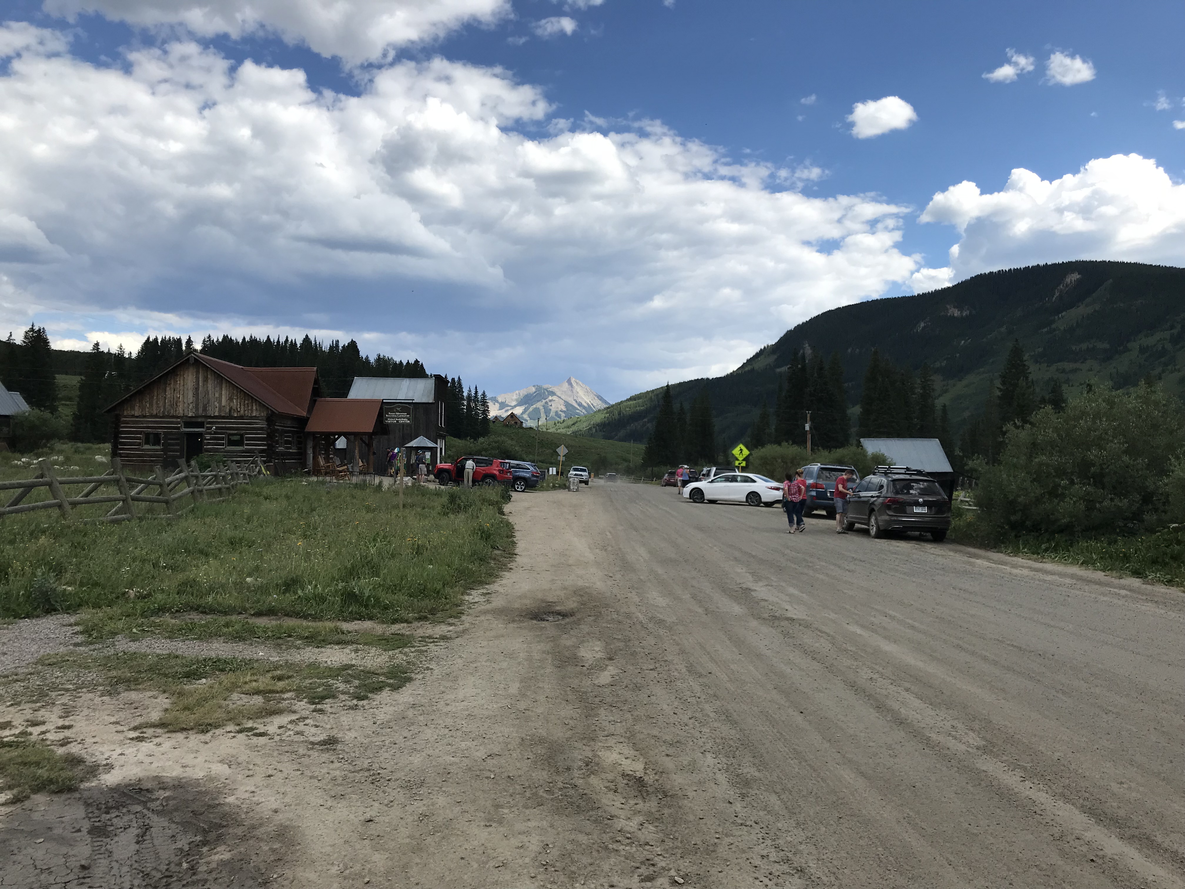 Modern cars line both sides of the dirt street. Old cabins sit in front of a rocky mountain far in the background