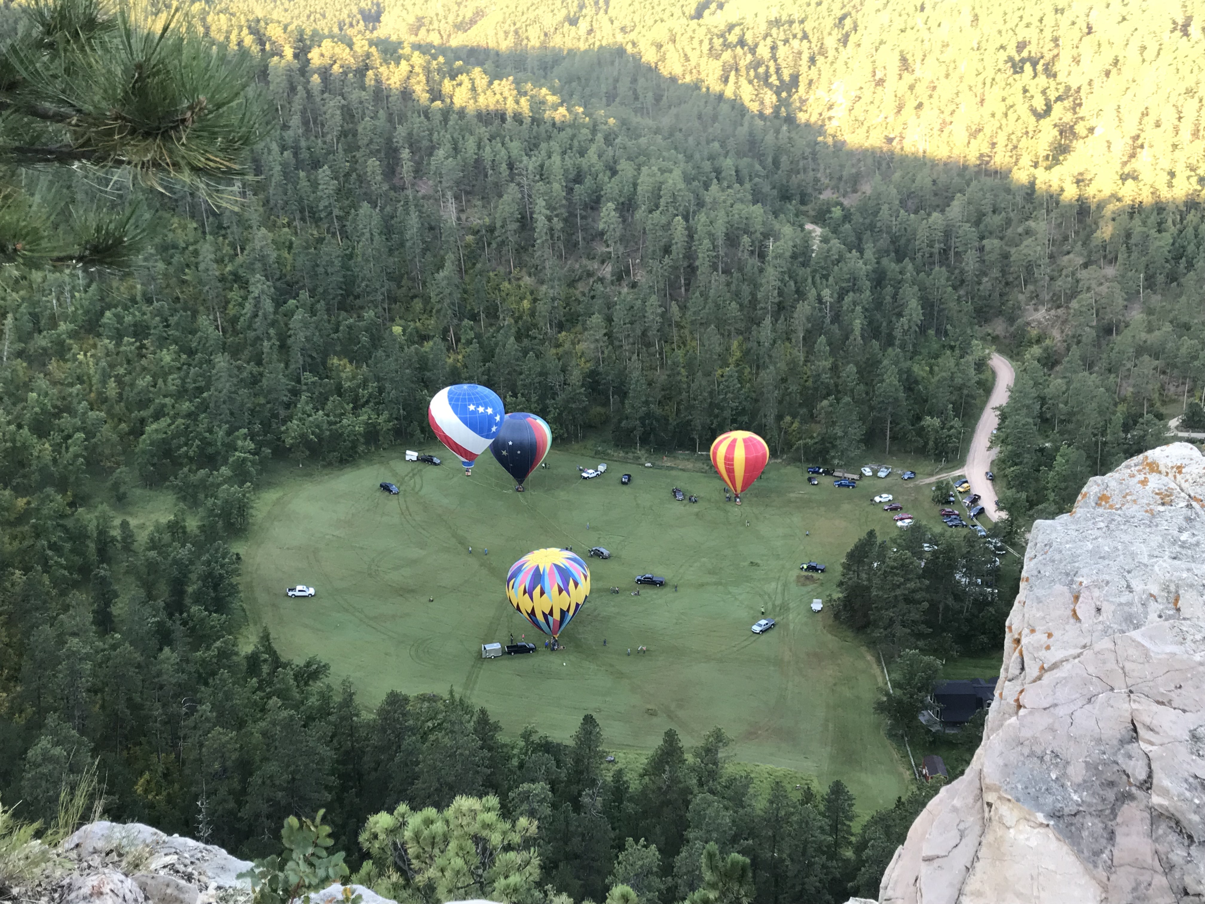 Top view of four colorful, hot air balloons floating near a grassy, valley floor surrounded by pine trees. A peace sign has been mowed into the grass. Cars and trucks also dot the valley floor.