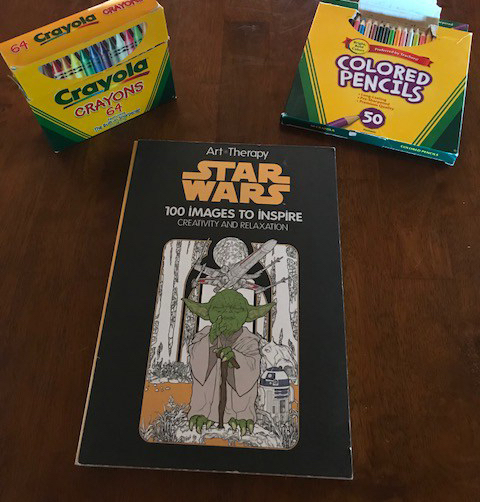 A box of crayons and colored pencils sit on a table. Between them is a Star Wars coloring book